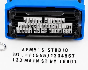 May include: A blue and black self-inking rubber stamp with a rectangular base. The stamp features raised white characters that read AEMY'S STUDIO, TEL: +1 (555) 1234567, and 123 MAIN ST NY 10001. A black ink impression of the same text is visible below.