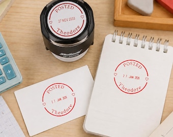 May include: A black self-inking stamp with a circular red ink impression reading POSTED, 27 NOV 2033, and Theodore with two small hearts. The stamp is shown next to a white card and a spiral notebook, both featuring the same red circular stamp design.