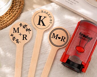 May include: Three light-colored wooden spoons with circular heads featuring black ink designs. One spoon shows M+R with leaf accents, one shows BAKED BY K, and one shows M heart R. A red Trodat Printy 46019 self-inking stamp sits to the right.