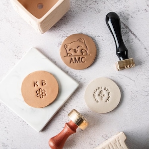 May include: An overhead shot of various craft stamps and materials. The image includes a wooden stamp with a black handle, round leather stamps with embossed designs, and a small ceramic planter. The stamps feature initials, a cat design, and the text "MADE BY ILANA."