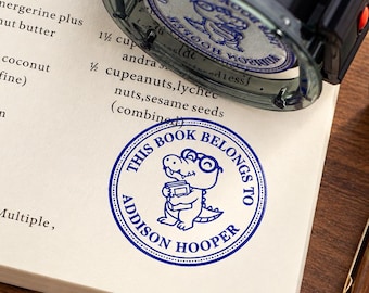 May include: A self-inking rubber stamp with a black casing, positioned above a cream-colored page. The stamp leaves a circular blue ink impression featuring a cartoon alligator wearing glasses and holding a book, with the text THIS BOOK BELONGS TO ADDISON HOOPER.