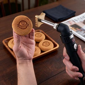 May include: A branding iron with a brass head is held near a round baked good with a logo. The baked good is held in a hand. Other baked goods with logos are in a wooden tray. The branding iron has a black handle.