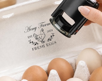 May include: A black self-inking rubber stamp positioned above an open egg carton. The stamp leaves a black ink impression on the carton lid featuring a chicken illustration, the text Hemy Farm, EST 2024, and the words FRESH EGGS.