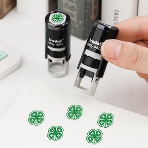 Op de afbeelding: Twee zwart-grijze Trodat Printy 4612 zelfinktende stempels. Eén stempel wordt rechtop gehouden, terwijl de andere op een oppervlak staat. Beide hebben een groen klavertjevier-icoon op de dop. Vijf groene klaverafdrukken zijn zichtbaar op het witte vlak.