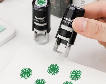 May include: Two black and gray Trodat Printy 4612 self-inking stamps. One stamp is held upright, while the other stands on a surface. Both feature a green four-leaf clover icon on the top cap. Five green clover imprints are visible on the white surface.