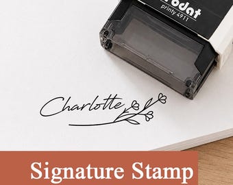 May include: A black and white Trodat Printy 4911 self-inking stamp positioned above a stamped impression on white paper. The impression features the name Charlotte in a cursive script font accompanied by a simple line-drawn floral branch design.