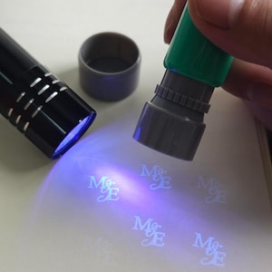 May include: A black UV light and a green and grey stamp are shown illuminating the letters "M&E" on a white surface. The stamp is held by a person. A grey cap is next to the UV light.