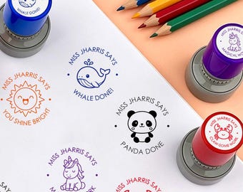 May include: A collection of round self-inking stamps with colorful caps and matching ink impressions on white paper. Designs include a whale with text 'MISS JHARRIS SAYS WHALE DONE!', a sun with 'YOU SHINE BRIGHT', a panda with 'PANDA DONE', a unicorn with 'MAGICAL WORK', and a crab with 'CLAW-SOME WORK'.