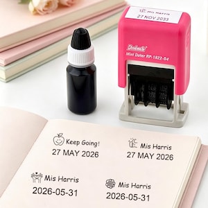 Peut inclure: Un tampon rose Deskmate Mini Dater RP-1822-04 avec un affichage de date, à côté d'un flacon d'encre noire. Le tampon affiche la date "27 NOV 2033". Un carnet affiche des dates et des phrases estampillées comme "Keep Going!" et "Mis Harris".