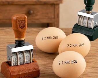 May include: A wooden date stamp with a chrome mechanism and a base engraved with the words PAR AVION, alongside three light brown eggs each stamped with the date 22 MAR 2026 in black ink, and a second green handled stamp in the background.
