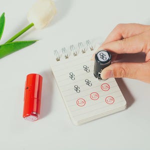 Op de afbeelding: Een rode en zwarte stempel wordt boven een klein notitieblok gehouden, met de afdruk van de stempel zichtbaar op de pagina. Er is ook een tweede rode stempel aanwezig. Het notitieblok heeft een spiraalbinding en is gelijnd. De stempelafdrukken zijn in zwarte en rode inkt.