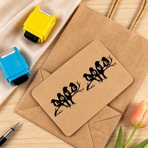 Könnte beinhalten: Eine rechteckige hellbraune Karte mit einer schwarzen Silhouettengrafik von zwei Vogelgruppen auf Ästen, wobei das Wort LOVE in die Vogelformen integriert ist. Die Karte liegt auf einer braunen Papiertüte neben einem gelben und einem blauen Stempel.