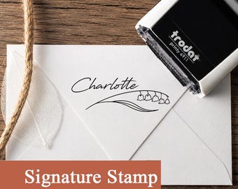 May include: A black and white Trodat Printy 4911 self-inking stamp resting on a white envelope. The stamp imprint features the name Charlotte in a cursive script above a line drawing of a lily of the valley flower with five small bell-shaped blooms.