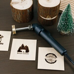 May include: A wood-burning tool with a black handle and a brass tip, next to several square wooden coasters with engraved designs. One coaster features a bear and trees, another says "Beare family."