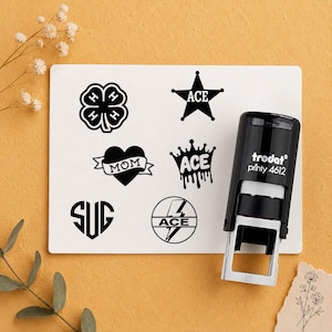 Op de afbeelding: Een zwarte Trodat Printy 4612 zelfinktende stempel naast een witte rechthoekige kaart met zes zwarte afdrukken: een klavertjevier met H's, een ster met ACE, een hart met MOM, een kroon met ACE, een hart met SUG en een bliksemschicht met ACE.