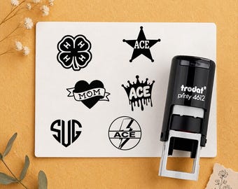 May include: A black Trodat Printy 4612 self-inking stamp next to a white rectangular card featuring six black printed designs: a four-leaf clover with H's, a star with ACE, a heart with MOM, a crown with ACE, a heart with SUG, and a lightning bolt with ACE.