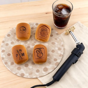 May include: Four small, golden-brown burger buns with various designs and text, including "LM", a burger graphic with "Bobo's Burgers", a fork and knife with "F M", and a peace sign. A glass of cola with ice and a branding iron are also present.