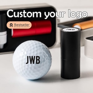 Puede incluir: Una pelota de golf blanca con las letras negras JWB impresas en la superficie, junto a un sello cilíndrico negro con las letras JWB en la parte superior. Al fondo, una caja metálica contiene un objeto cilíndrico rojo y el texto Custom your logo.