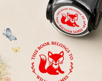 May include: A black self-inking stamp with a circular impression featuring a red fox illustration, a leafy wreath border, and the text "THIS BOOK BELONGS TO WILLIAM JAMES" printed in a bold red font, shown with a matching red stamped imprint.
