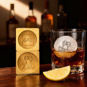 Op de afbeelding: Twee gouden vierkante ijsblokjesstempels, één met "2AS" en de andere met "A&M" gegraveerd. Een glas whisky met een grote ijsbol met een wijnglasontwerp en een citroenpartje.