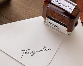 May include: A self-inking stamp with a dark wood-grain finish and a clear base, labeled Trodat Printy T926. The top displays the text Thesignature in a script font, which is also stamped in black ink onto a white envelope below the device.