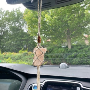 Może przedstawiać: Wisząca ozdoba do samochodu z jasno-różowym kryształem owiniętym w siatkę makramy, zawieszona na kremowym sznurku. Drewniana koralik z rzeźbionymi detalami znajduje się nad kryształem. Ozdoba wisi na lusterku wstecznym w samochodzie.