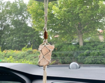 Puede incluir: Un adorno colgante para coche con un cristal rosa claro envuelto en una red de macramé, suspendido de un cordón color crema. Una cuenta de madera con detalles tallados se coloca encima del cristal. El adorno cuelga del espejo retrovisor de un coche.