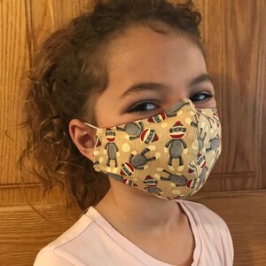 May include: A child wearing a beige face mask with a pattern of gray sock monkeys. The mask has a white elastic band.