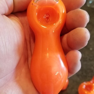 May include: An orange glass pipe with a rounded bowl and a long, curved stem. The pipe is held in a hand, and other similar pipes are visible in the background. The glass has a glossy finish.
