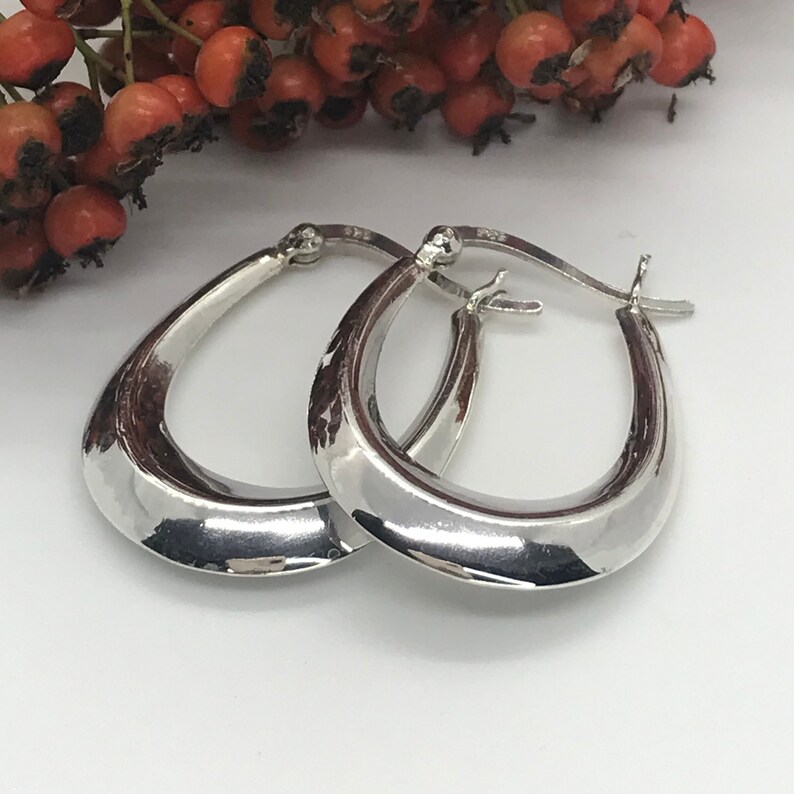 May include: A pair of silver hoop earrings with a smooth, curved design. The earrings are oval shaped and have a polished finish.