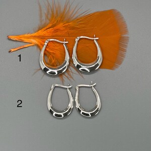 May include: Two pairs of silver hoop earrings. The top pair is larger than the bottom pair. The earrings are oval shaped and have a smooth finish.