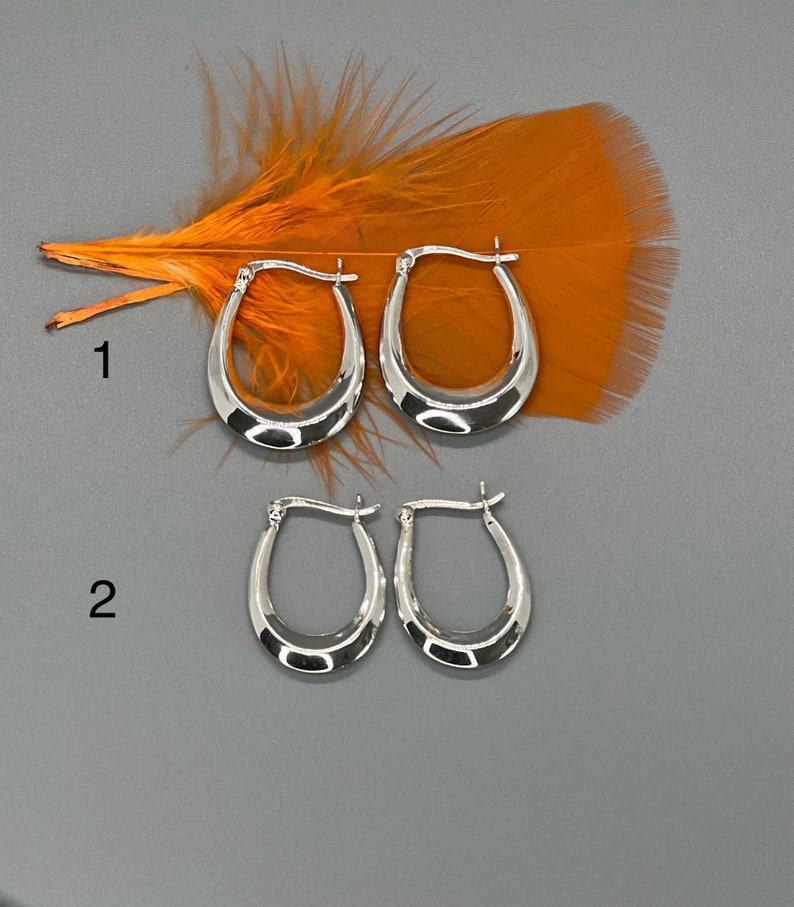 May include: Two pairs of silver hoop earrings. The top pair is larger than the bottom pair. The earrings are oval shaped and have a smooth finish.