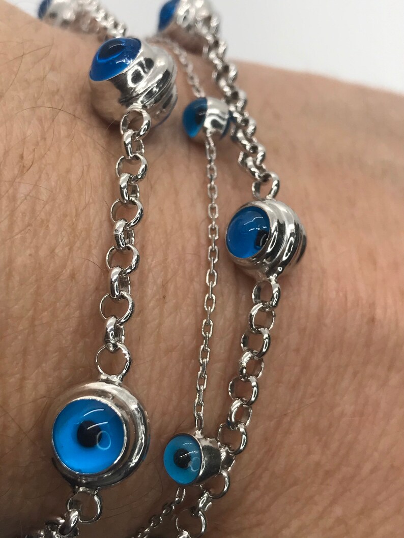 May include: A silver chain bracelet with multiple blue evil eye charms. The charms are round and have a silver setting.