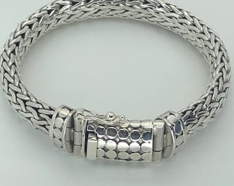 May include: A silver bracelet with a woven chain design. The bracelet features a secure clasp with a series of small, circular indentations. The bracelet is a piece of jewelry.
