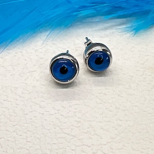 Può includere: Un paio di orecchini a perno d'argento con ciondoli a forma di occhio blu maligno. Gli orecchini sono su una superficie bianca con una piuma blu sullo sfondo.