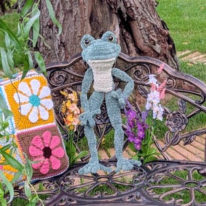 May include: A crochet frog stands on a decorative metal bench with floral accents. A colorful crochet pillow with flower designs rests on the bench. The scene is set outdoors with a tree and green grass.