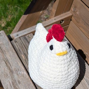 May include: A white crochet chicken with a red comb, black eyes, and a yellow beak. The soft toy is sitting on a wooden bench. This handmade item is ideal for farmhouse decor or as a unique gift idea.