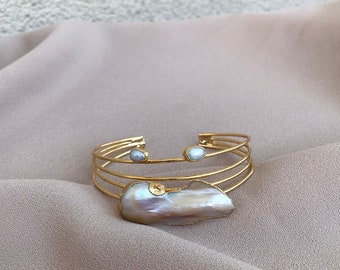 May include: A gold bangle bracelet with a large, iridescent, pearlescent shell charm and two smaller pearl accents. The bracelet is on a light brown fabric background.