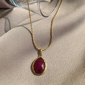Könnte beinhalten: Eine goldene Kettenkette mit einem tropfenförmigen Anhänger mit einem tief roten Edelstein.