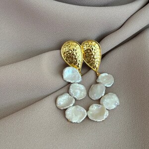 Può includere: Orecchini dorati con un design a goccia testurizzato e un gruppo di perle barocche bianche.