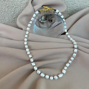 Könnte beinhalten: Eine weiße Perlenkette mit einem goldenen Verschluss an einer schwarzen Schnur. Die Kette liegt auf einem hellbraunen Stoff.