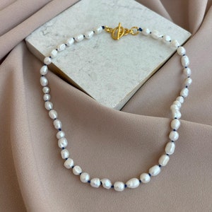 Könnte beinhalten: Eine weiße Perlenkette mit einem goldenen Verschluss und einer blauen Schnur. Die Kette liegt auf einem hellbraunen Stoffhintergrund.