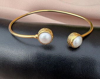 Puede incluir: Una pulsera abierta de tono dorado con dos cuentas redondas blancas similares a perlas en cada extremo. Los engastes de las perlas tienen una superficie texturizada y orgánica, y la banda es un alambre de metal dorado, fino, liso y circular.