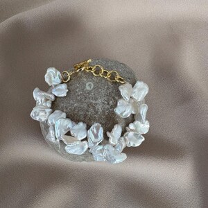 May include: A delicate bracelet featuring irregular, white, petal-shaped pearls. The pearls are strung together with a gold-colored chain and clasp. The bracelet is displayed on a gray, textured stone, set against a neutral background.