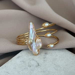 May include: A gold-toned cuff bracelet featuring a large, iridescent baroque pearl as a focal point. The bracelet has a spiral design with three smaller, oval-shaped pearls. The bracelet is displayed on a neutral background.