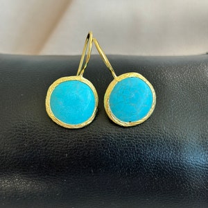 Puede incluir: Un par de pendientes redondos con piedras de color turquesa brillante engastadas en marcos de metal dorado texturizado. Cada pendiente cuelga de un gancho de alambre dorado curvado, mostrando una superficie azul vibrante y lisa.