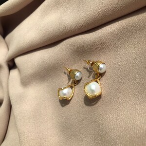 May include: A pair of gold-colored earrings with a pearl accent. The earrings have a post back and a unique design, with a small pearl at the top and a larger, irregularly shaped pearl suspended below. The earrings are set against a neutral background.