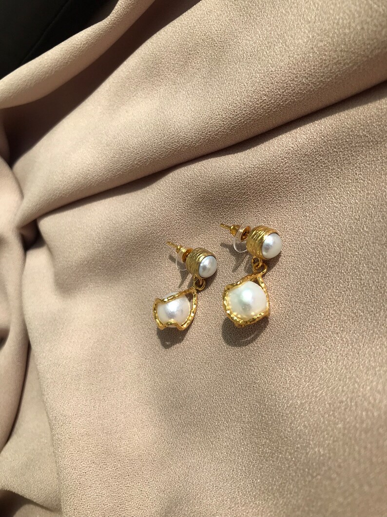 May include: A pair of gold-colored earrings with a pearl accent. The earrings have a post back and a unique design, with a small pearl at the top and a larger, irregularly shaped pearl suspended below. The earrings are set against a neutral background.