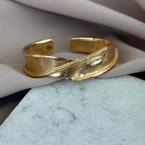 May include: A gold-colored cuff bracelet with a textured, folded design. The open-ended bracelet sits on a marble surface and a draped fabric background. The jewelry piece has a modern, minimalist aesthetic.