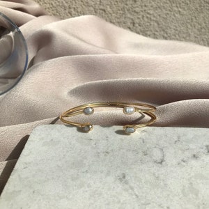 May include: A gold-toned cuff bracelet with four oval, light-colored stones. The bracelet has a minimalist design with a double-banded structure. The stones are evenly spaced along the top of the bracelet.